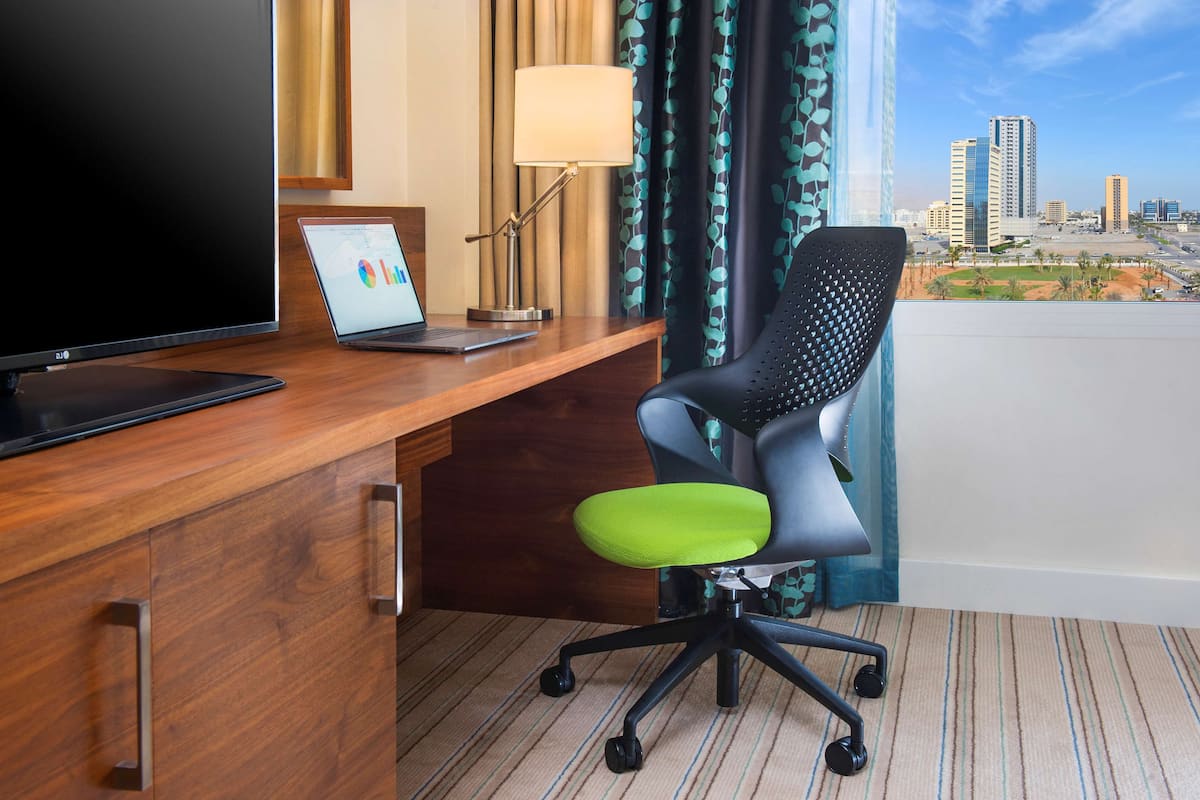 A hotel room with a wooden desk, a laptop, a television, a lamp, and a chair with a green cushion.