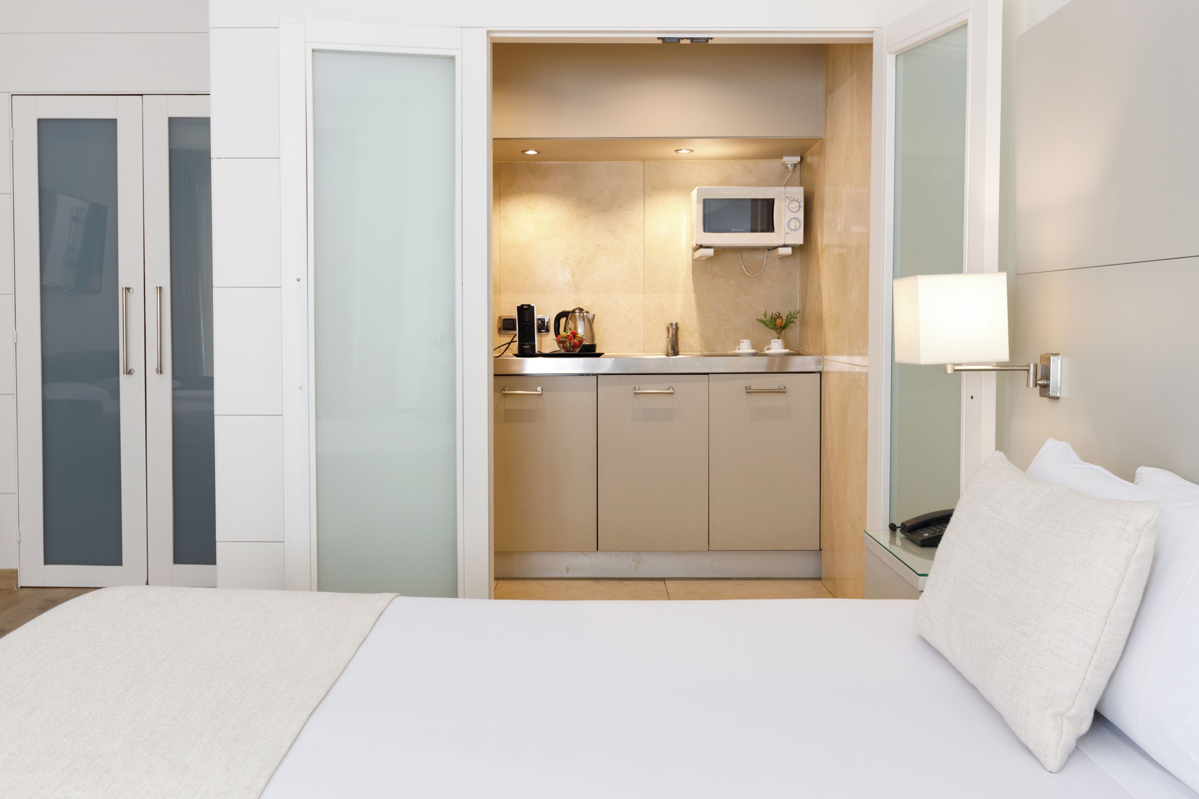 A bedroom with a bed, a nightstand, a bedside table, a wall-mounted lamp, and a sliding door leading to a kitchenette with a microwave, a coffee maker, and a small plant.