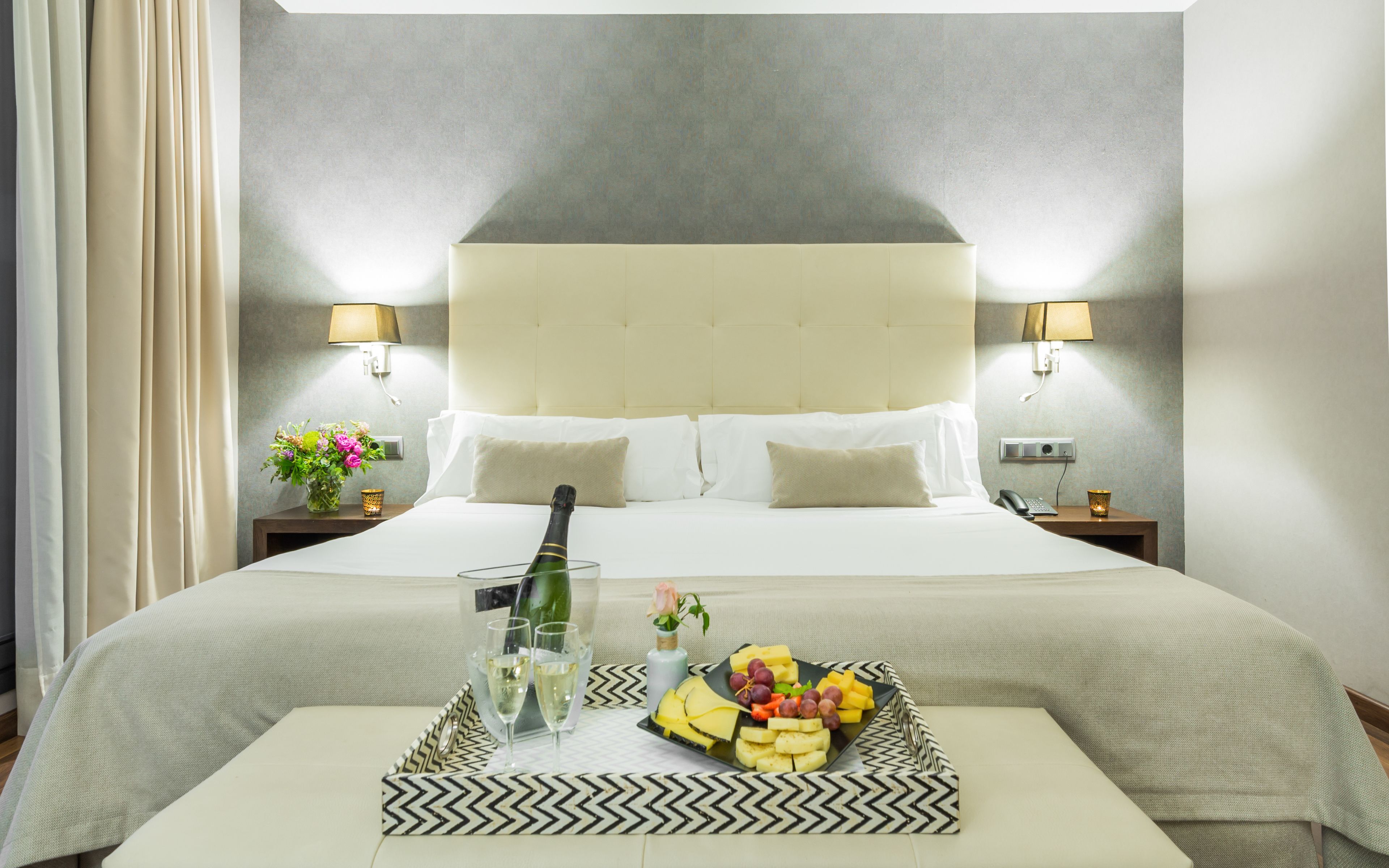 A hotel room with a large bed, two bedside lamps, a nightstand with a phone and a candle, and a tray with food and drinks.