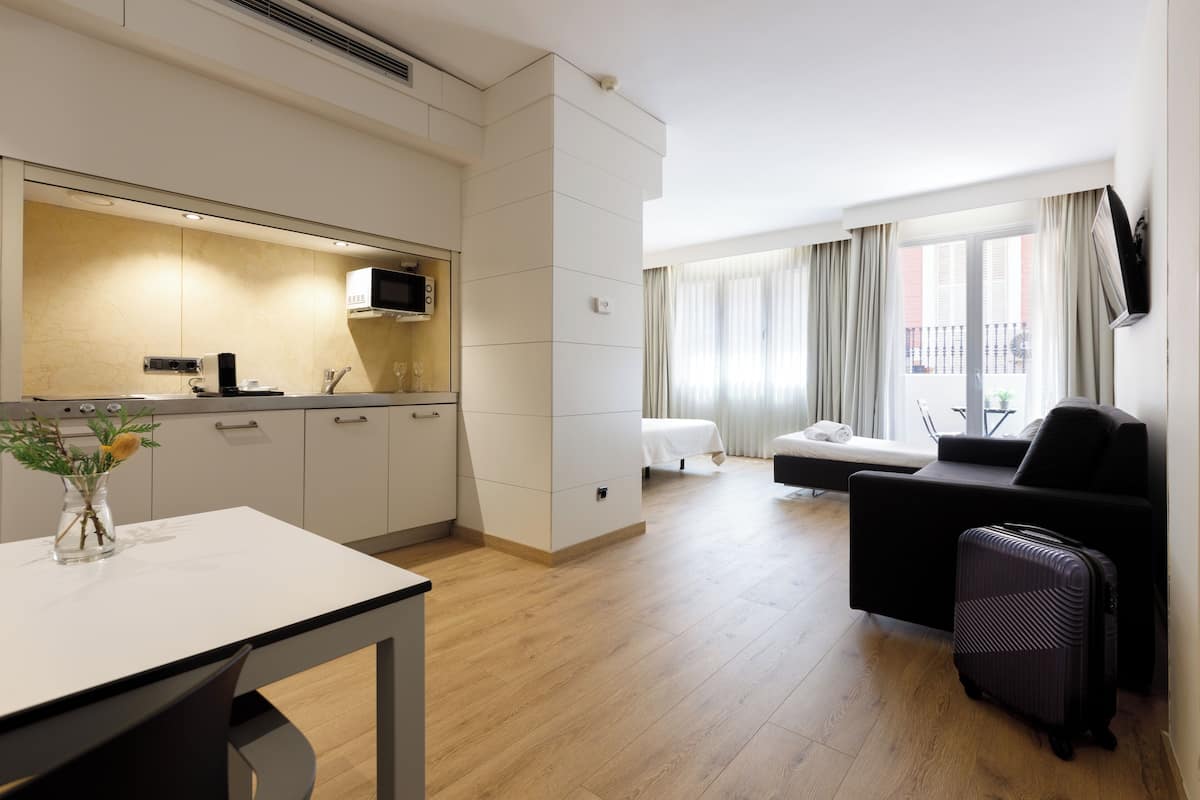 Une chambre d’hôtel moderne dotée d’une kitchenette, d’un coin repas, d’un espace de vie avec un canapé et une télévision, ainsi que d’un balcon agrémenté de rideaux.