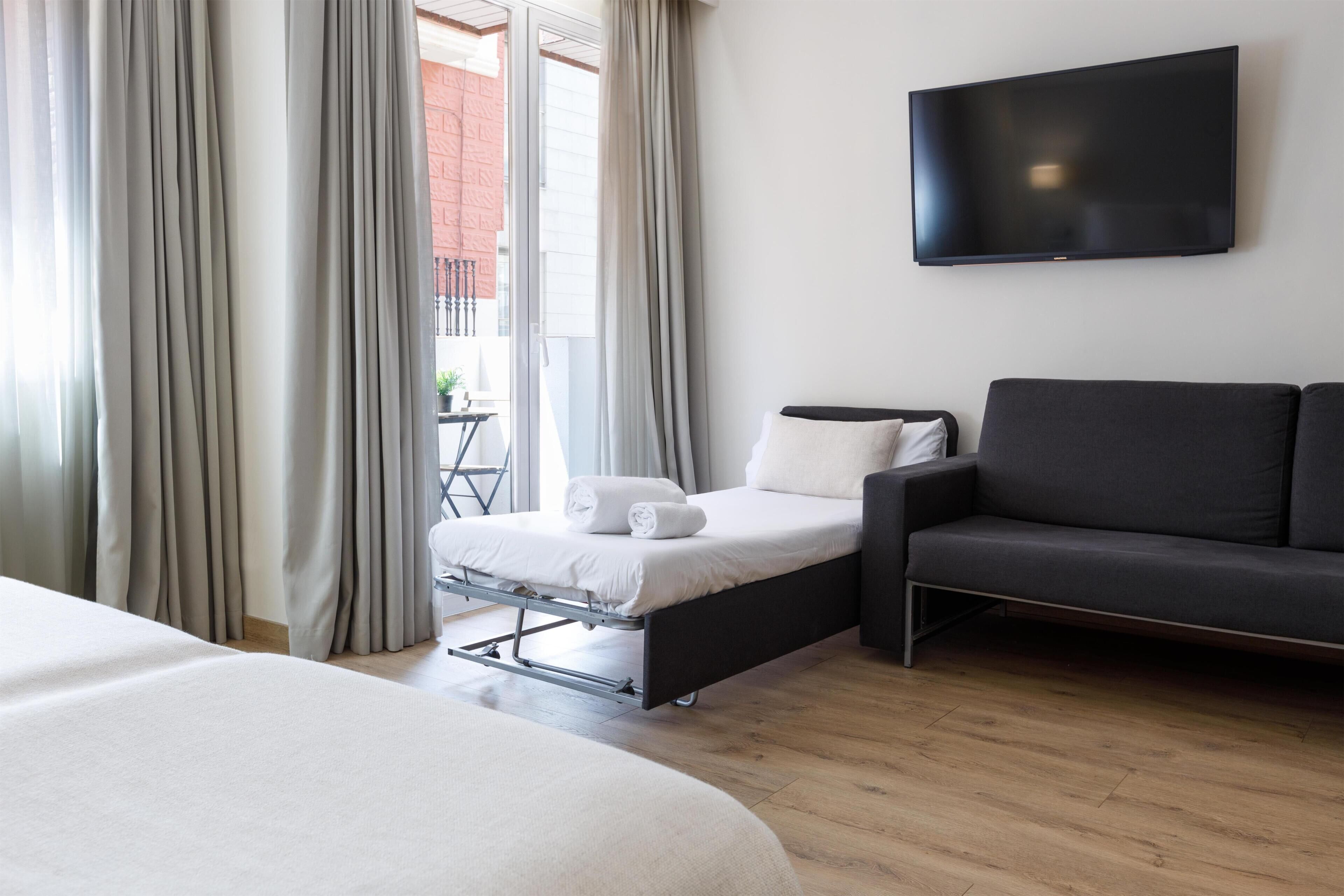 Ein Hotelzimmer mit einem Bett, einem Sofa, einem an der Wand montierten Fernseher und einem Fenster mit Vorhängen.