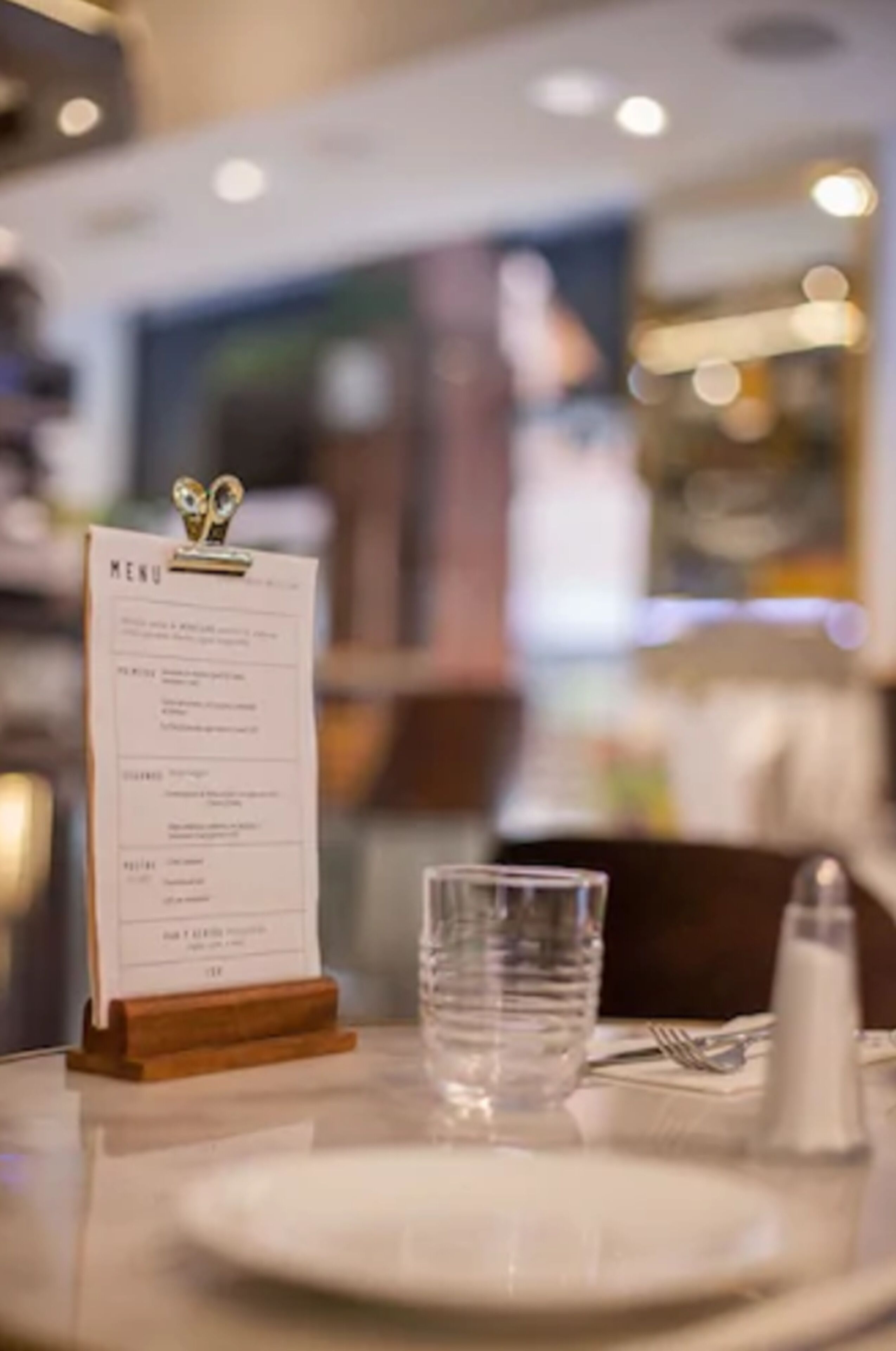 A restaurant setting with a menu on a wooden stand, a glass of water, and a plate on the table.