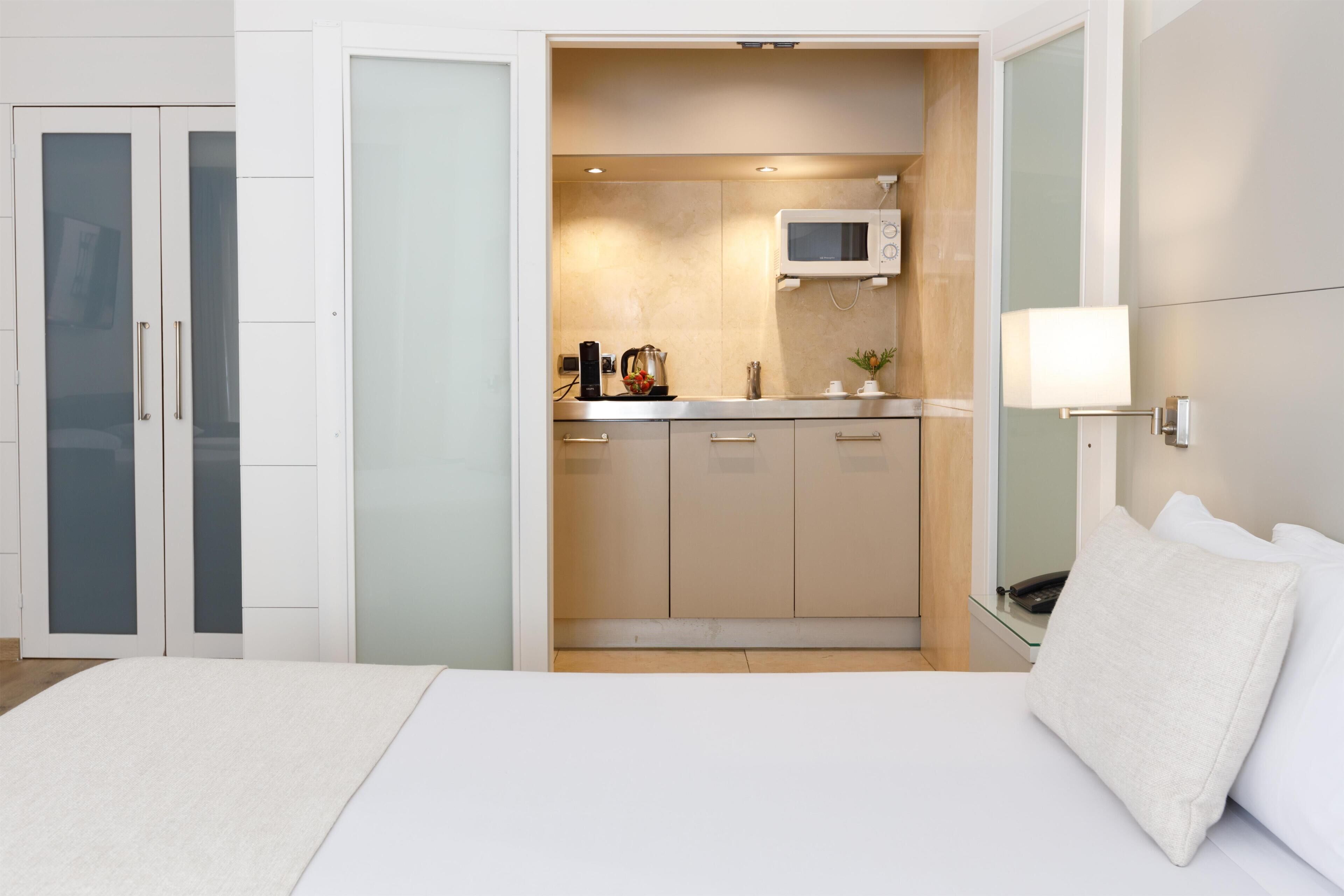 A bedroom with a bed, a nightstand, a bedside table, a wall-mounted TV, and a sliding door leading to a kitchenette with a microwave, a kettle, and a coffee maker.
