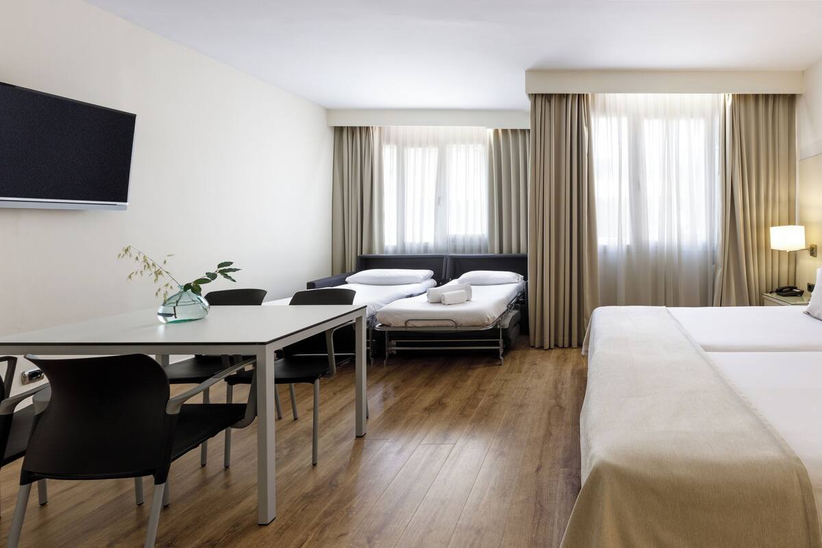 Une chambre d’hôtel comprenant un lit, un bureau avec des chaises, un téléviseur fixé au mur et une fenêtre avec des rideaux.
