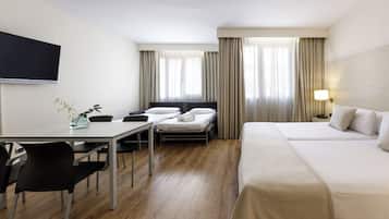 A hotel room with a bed, a desk with chairs, a TV mounted on the wall, and a window with curtains.