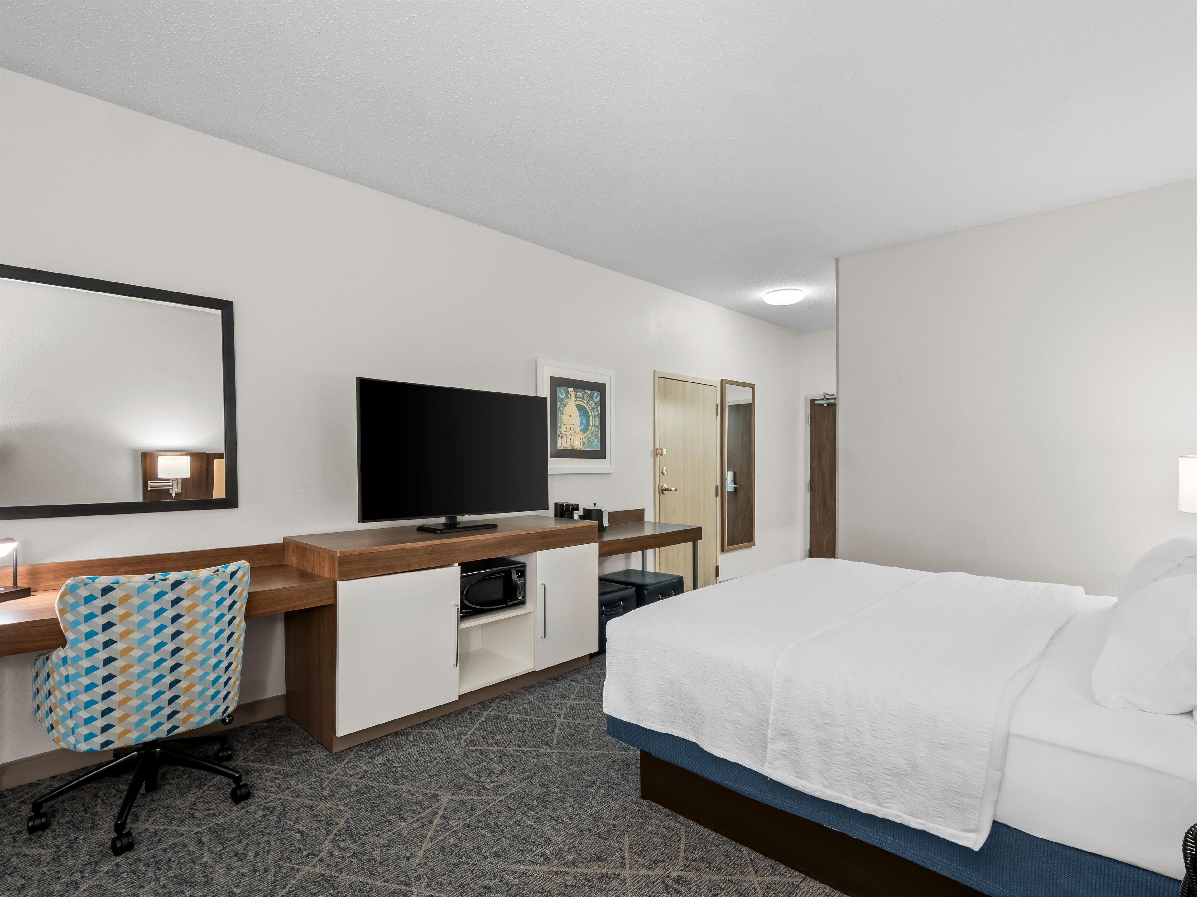 A hotel room with a bed, a desk with a chair, a television, a wardrobe, and a mirror.