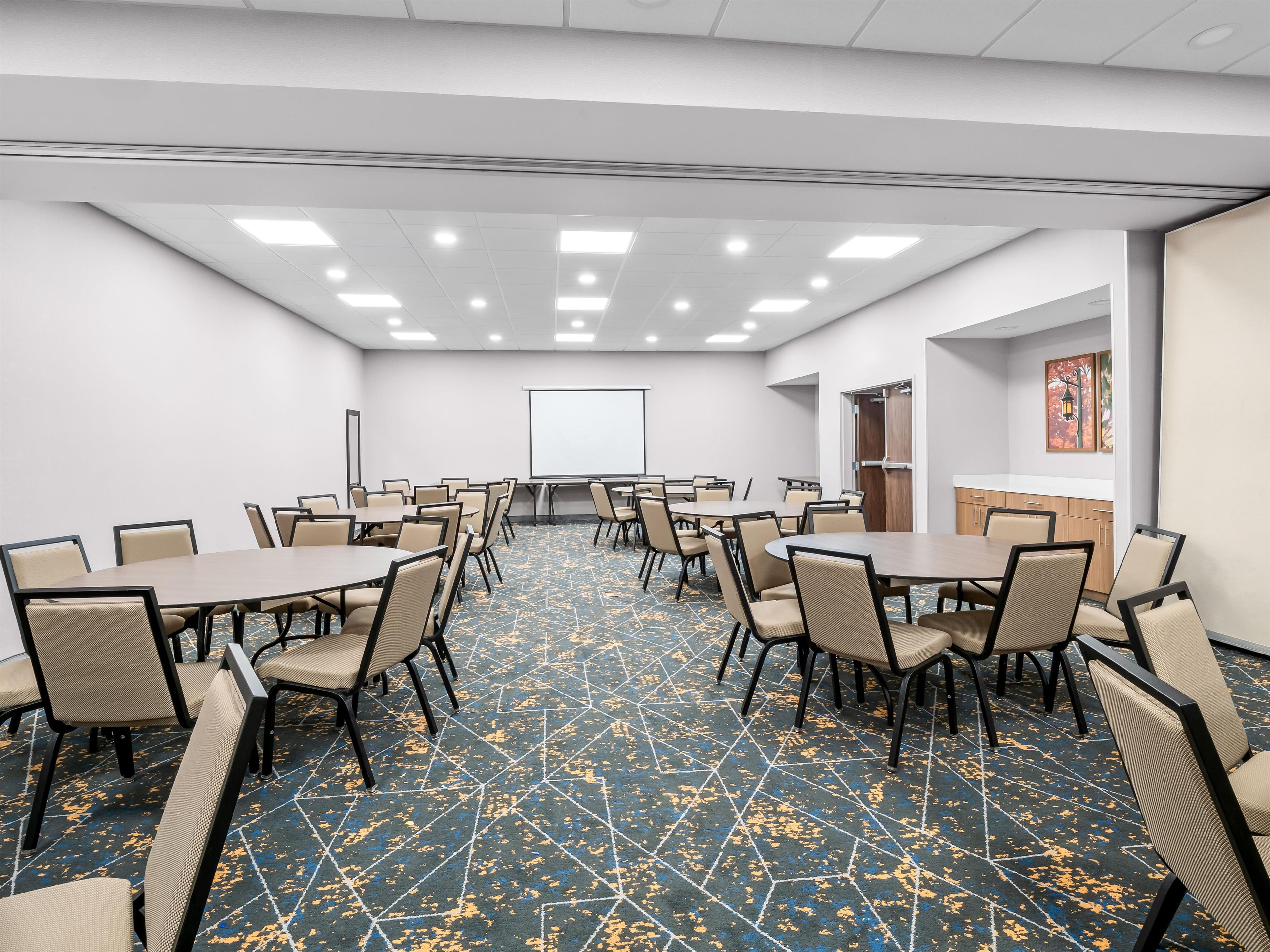 A conference room with multiple tables and chairs, a projector screen, and a patterned carpet.