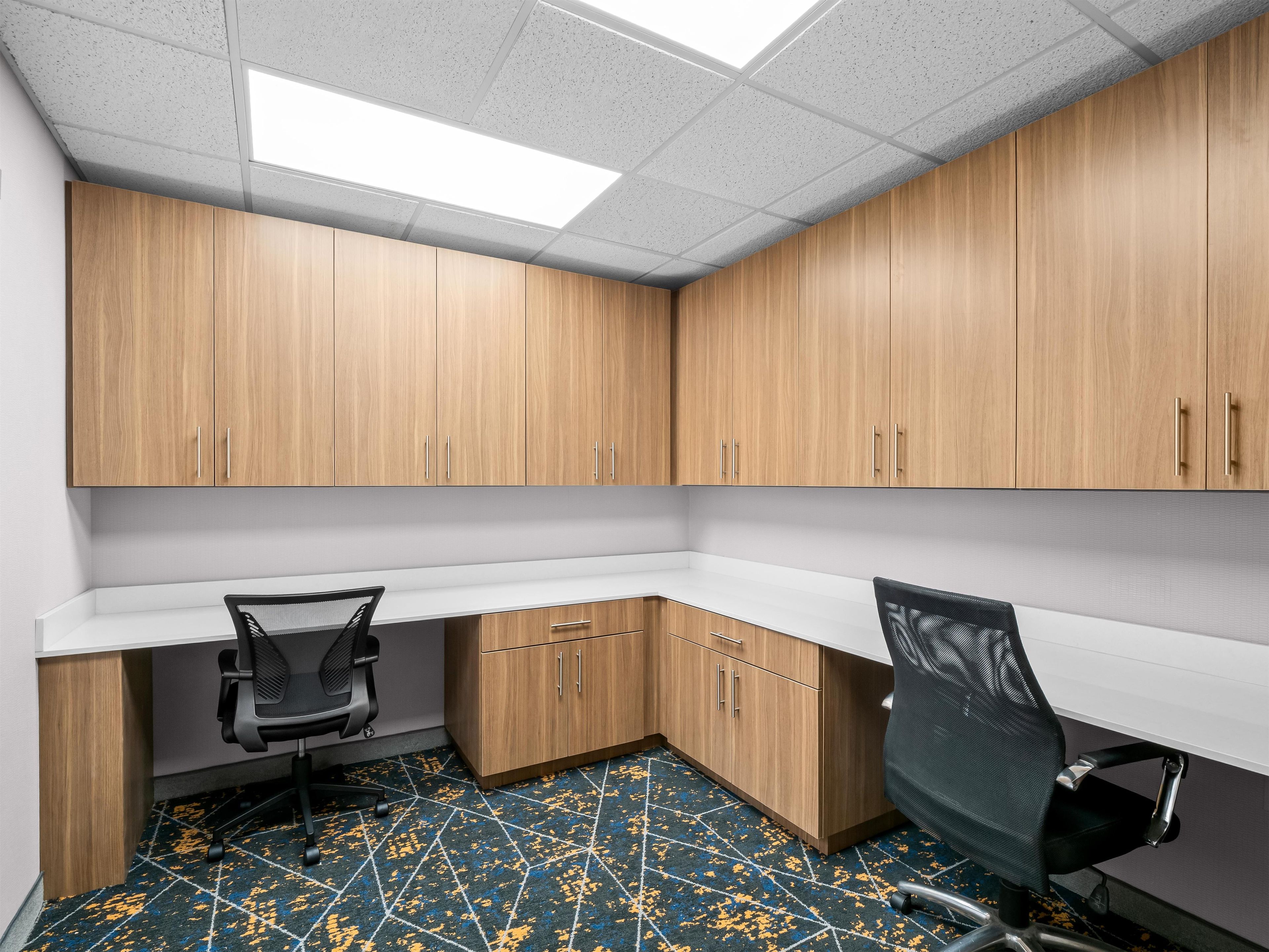 A room with wooden cabinets, two office chairs, a white desk, and a patterned floor.