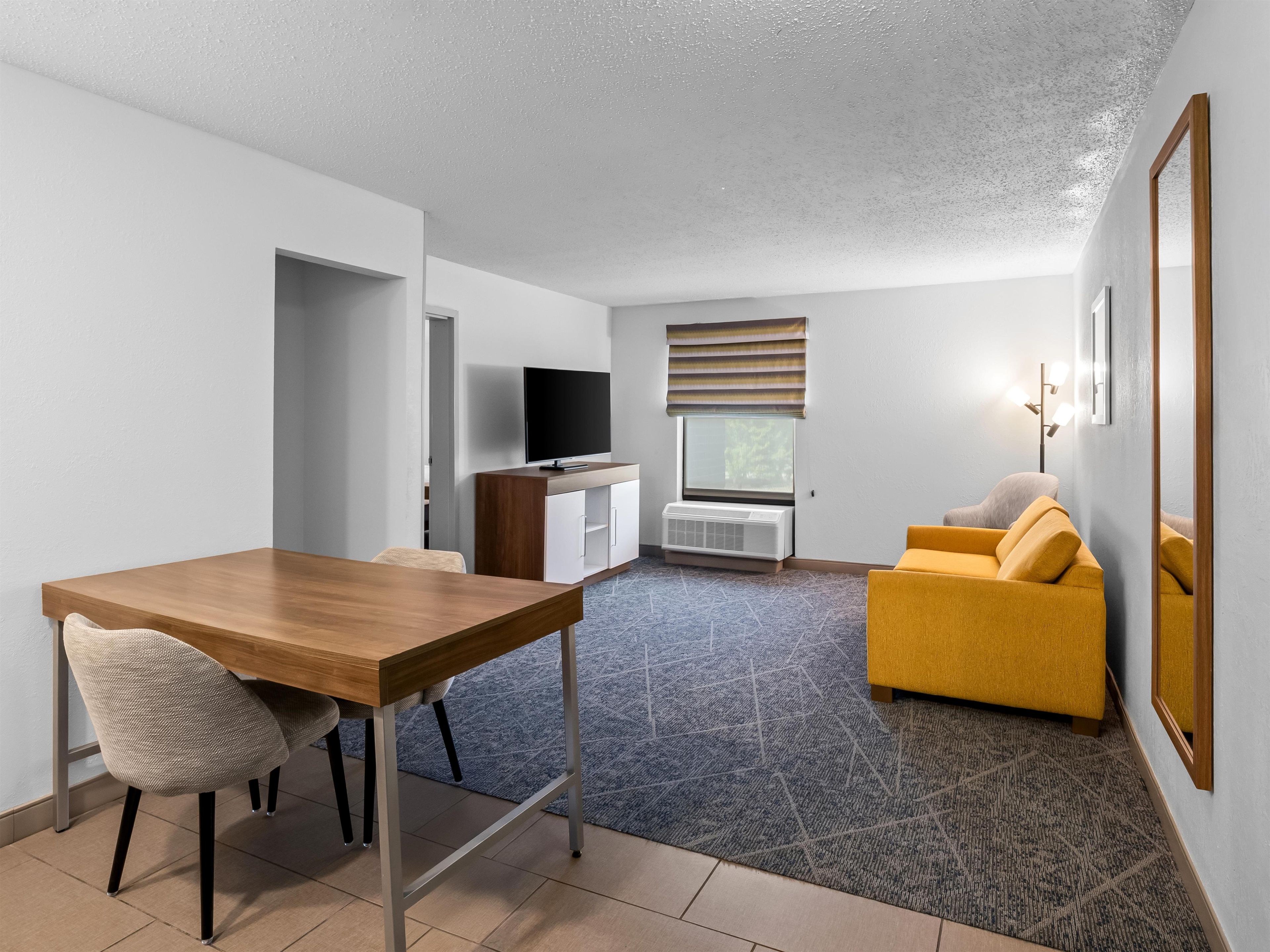 Ein modernes Hotelzimmer mit einem Holztisch, einer Couch, einem Fernseher und einem Fenster mit Jalousien.