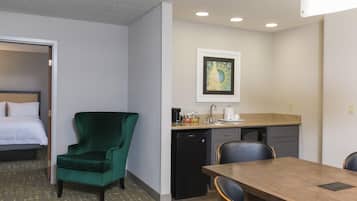 A hotel room with a bed, a green armchair, a dining table, a kitchen area, and a framed picture on the wall.