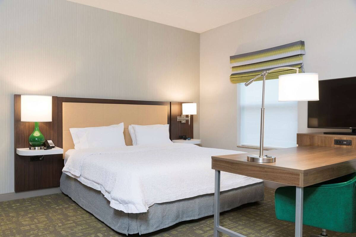 Quarto de hotel com cama, luminárias de cabeceira, escrivaninha, televisão e uma cadeira verde.