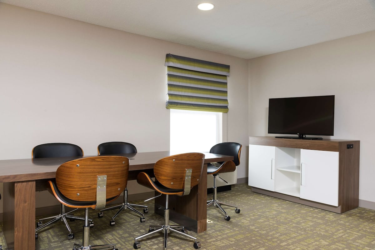 A conference room with a wooden table, four chairs, a television on a cabinet, and a window with blinds.