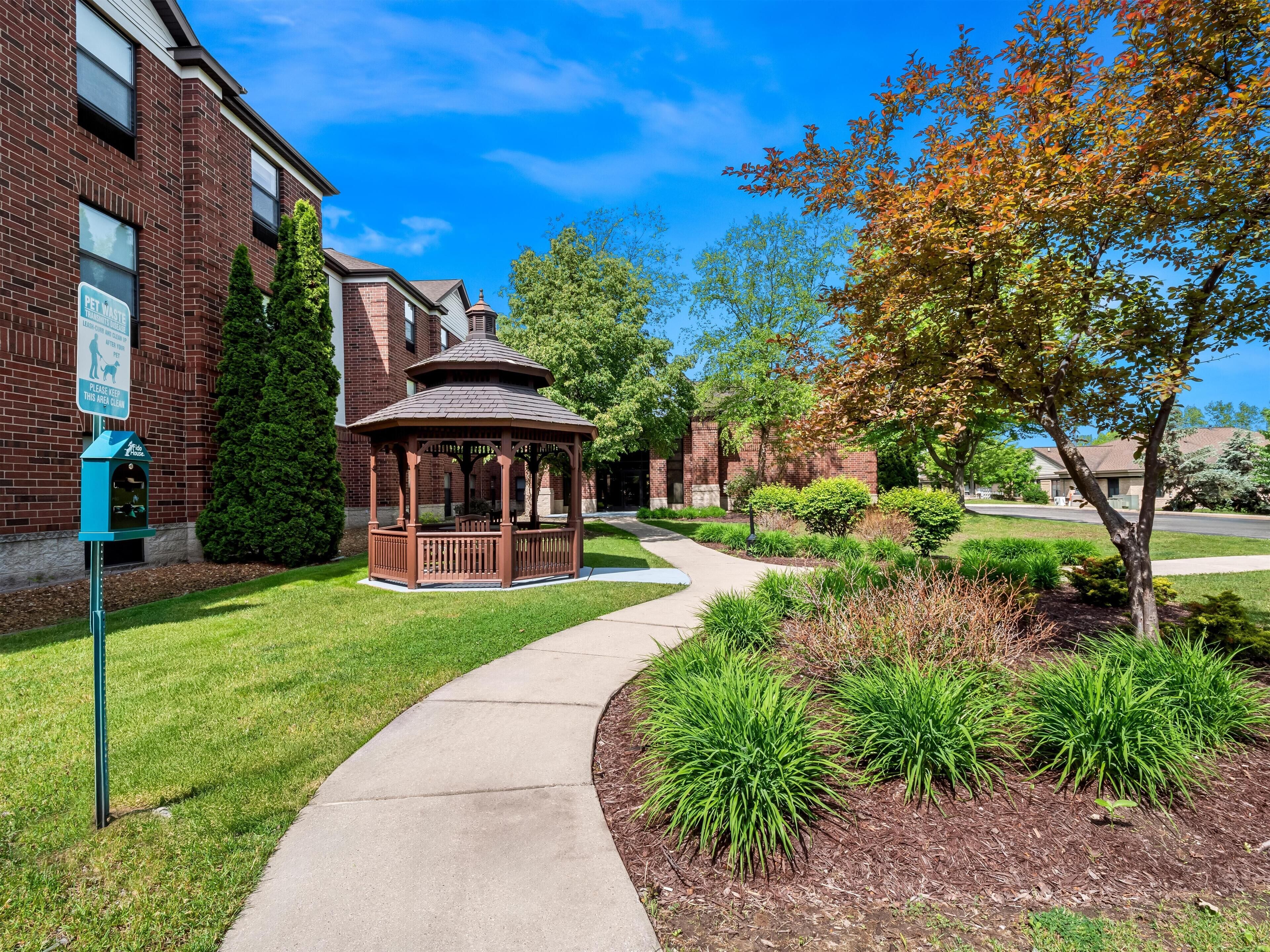 A gazebo in a landscaped area with a brick building in the background.