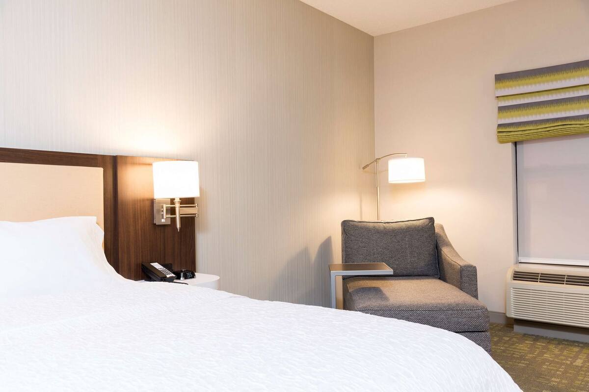 Quarto de hotel com uma cama, uma cadeira, uma mesa de cabeceira com abajur, um relógio de parede e uma janela com persianas.