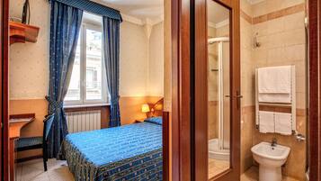 Ein Hotelzimmer mit Bett, Schreibtisch, einem Fenster mit Vorhängen, einem Badezimmer mit Dusche, Waschbecken und Spiegel.
