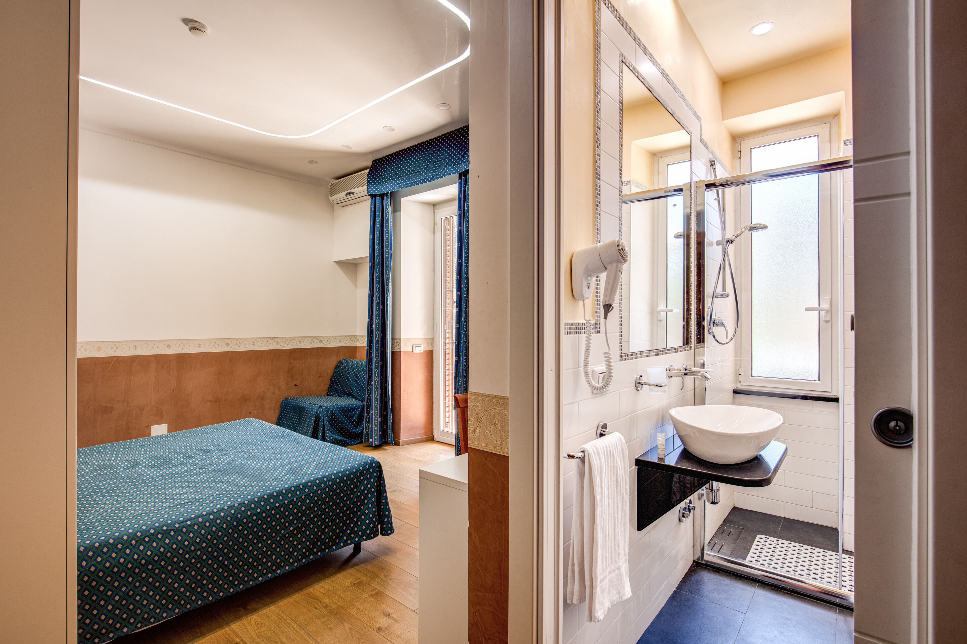Habitación de hotel con cama, un pequeño espacio para sentarse, baño con lavamanos, espejo y ducha.