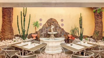 Um pátio externo com uma fonte de pedra, plantas de cactos e uma área de estar com cadeiras e mesas de metal.