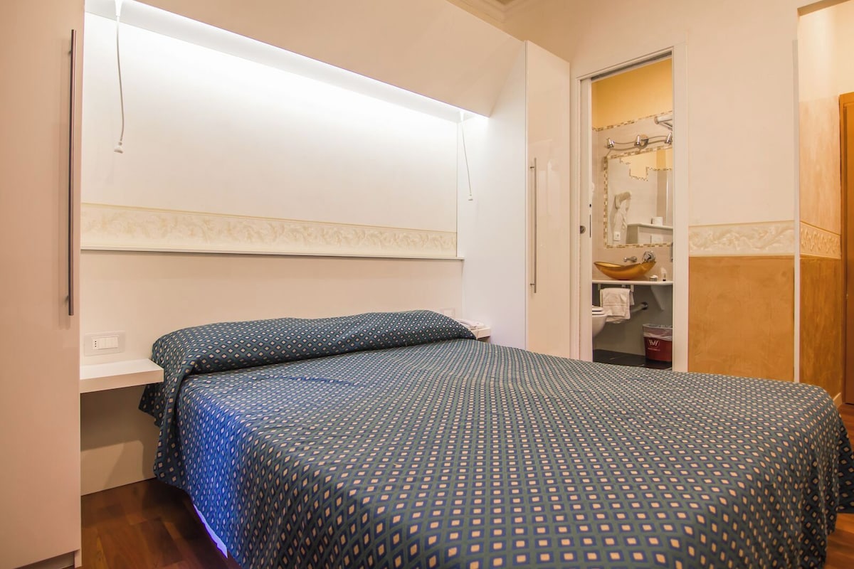 Un dormitorio con cama, mesita de noche, estante en la pared y puerta que da al baño.