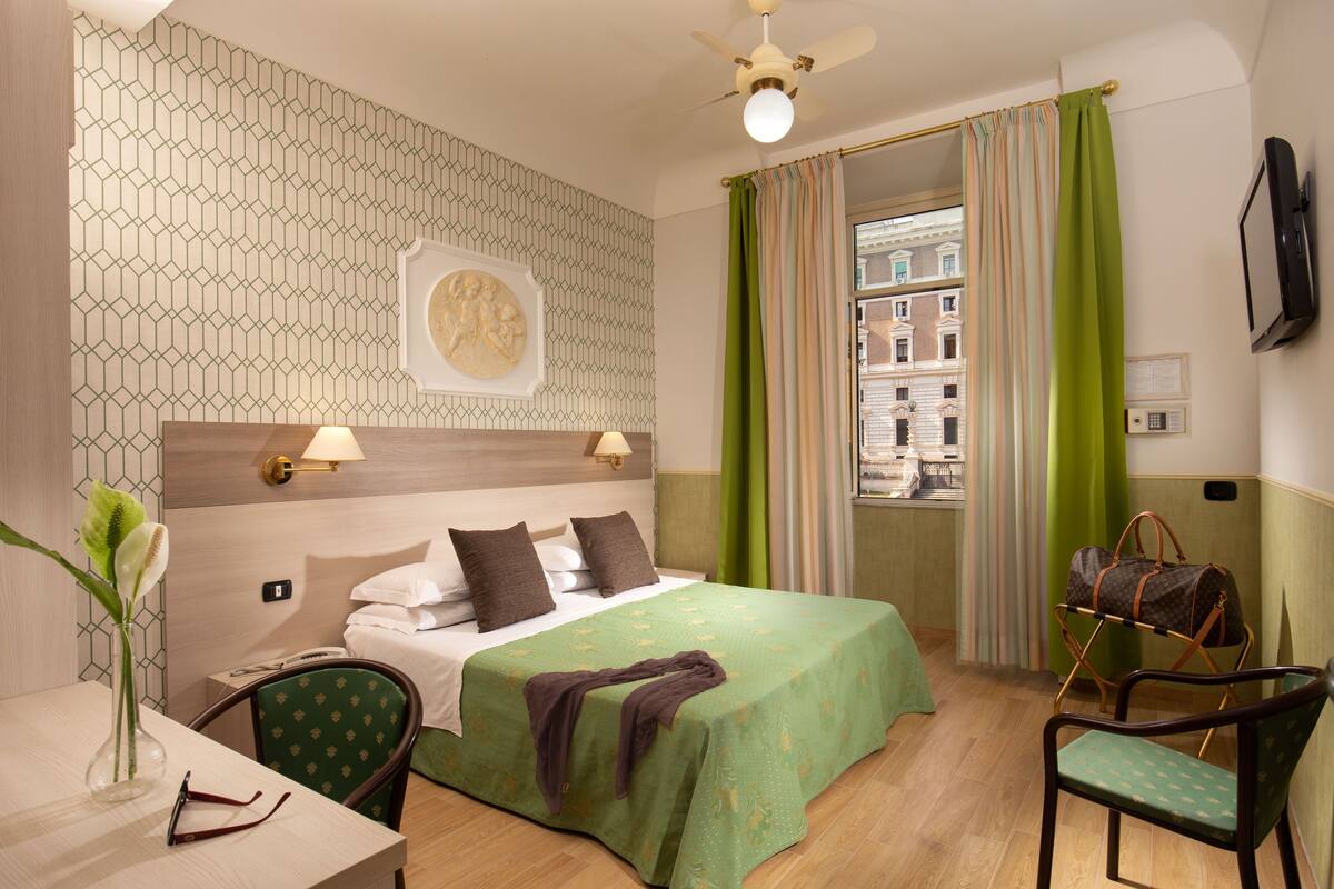 A hotel room with a bed, a desk with a chair, a vase with flowers, a lamp, and a view of buildings through a window with curtains.