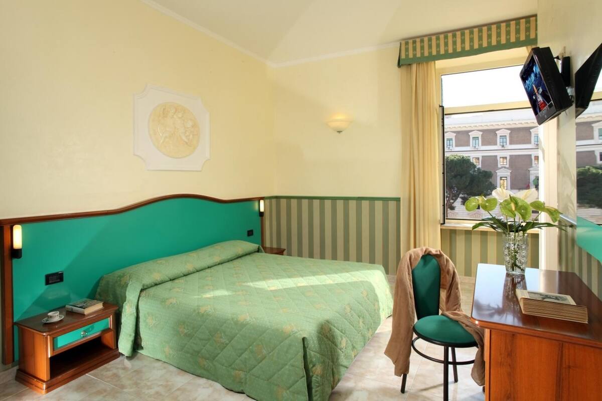 A hotel room with a bed, a desk, a chair, a TV, and a window with a view of buildings.