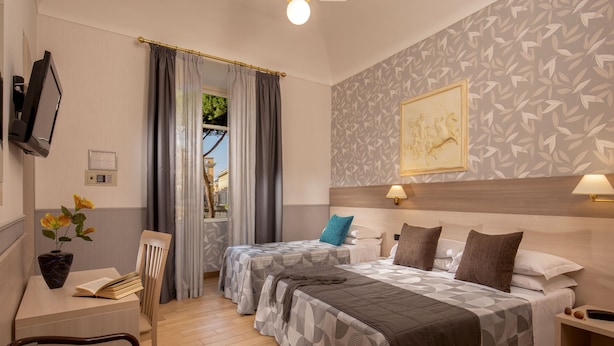 A hotel room with two beds, a desk with a chair, a TV, a window with curtains, and a patterned wall. - Hotel Dorica (Rome)