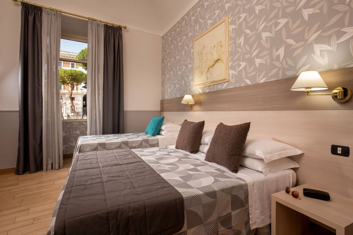 A hotel room with two beds, a window with curtains, a wall-mounted lamp, and a framed picture.