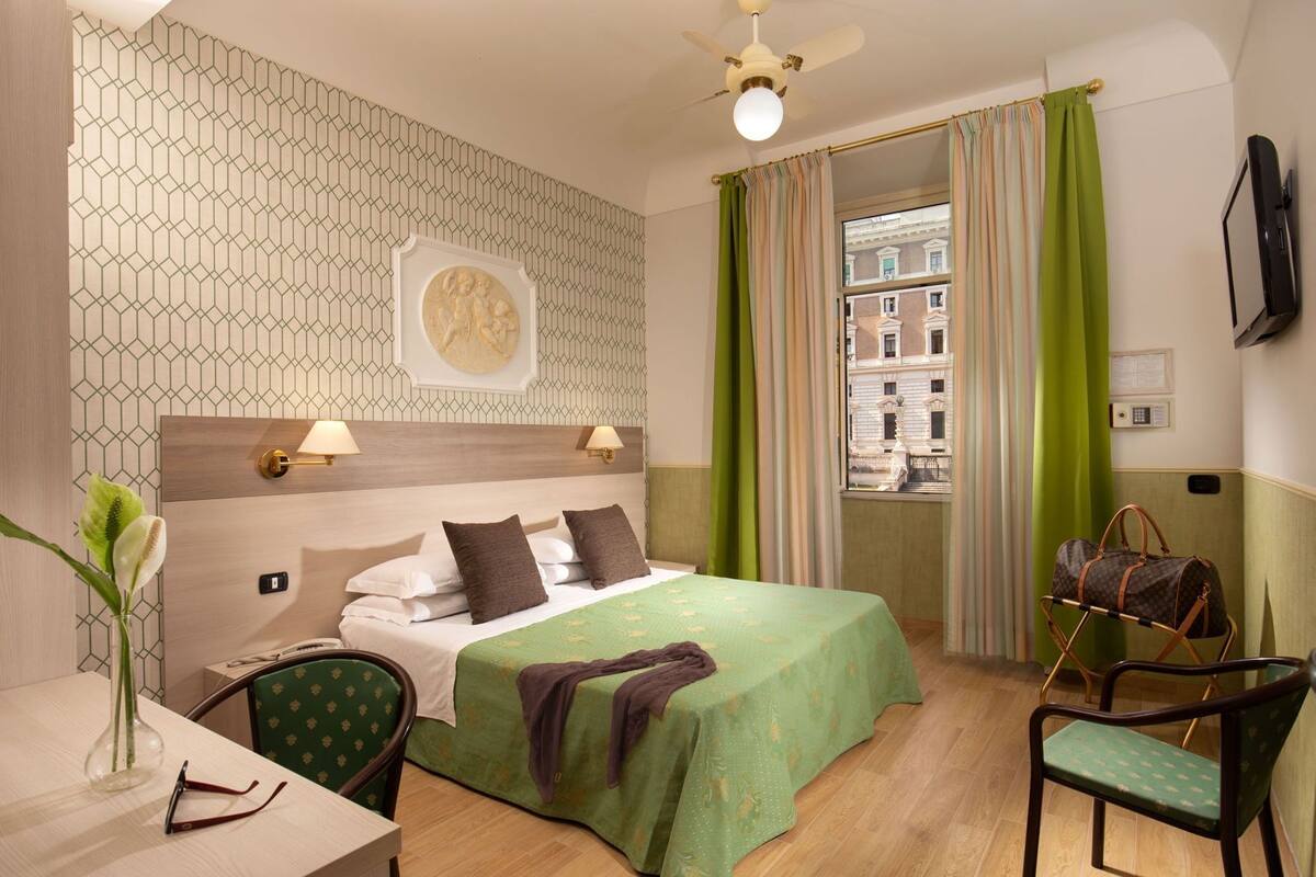 A hotel room with a bed, a desk, a chair, a TV, and a view of buildings.