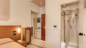 Quarto de hotel com cabeceira de madeira, telefone ao lado da cama, mesa de cabeceira pequena, banheiro com chuveiro em cabine de vidro e espelho.