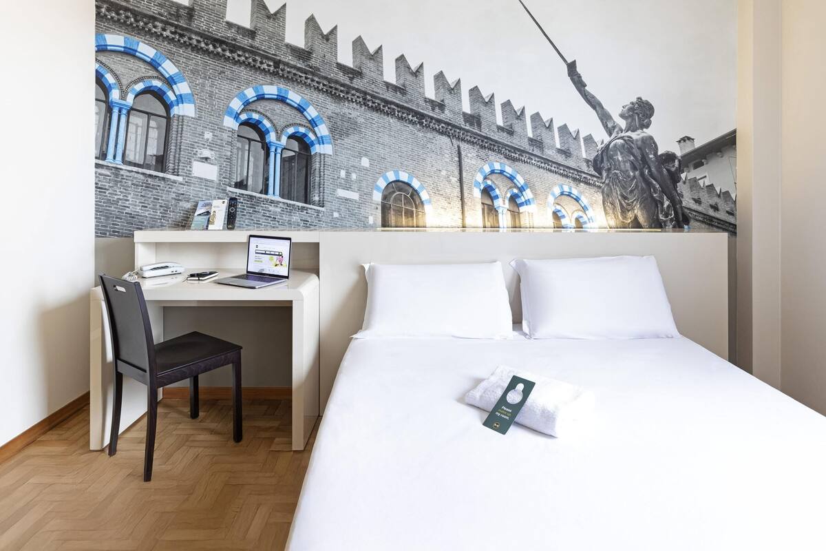A hotel room with a bed, a desk, and a chair. There is a mural on the wall depicting a castle and a statue.