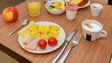 Una tavola per la colazione con uova strapazzate, prosciutto, pomodori, una ciotola di frutta, un croissant, un bicchiere di succo d'arancia, una tazza di caffè e un piatto con una mela.
