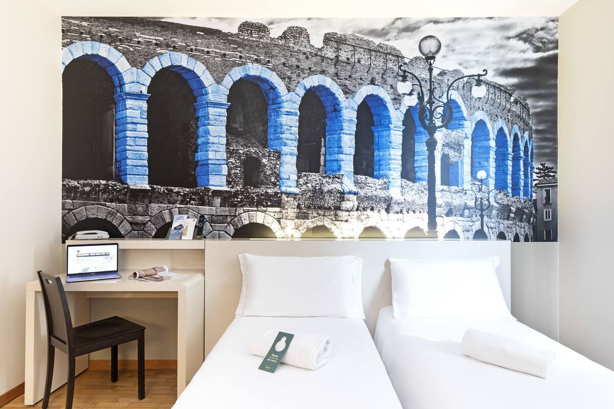 A hotel room with a bed, a desk with a laptop, and a large wall mural of blue arches.