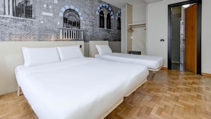 A hotel room with two beds, wooden parquet flooring, and a brick wall with arched windows. - B&B Hotel Verona (Verona)