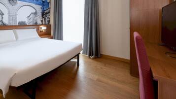 Ein Hotelzimmer mit einem großen Bett, Holzboden, einem Schreibtisch mit Stuhl, einem Fernseher und einem Wandgemälde.