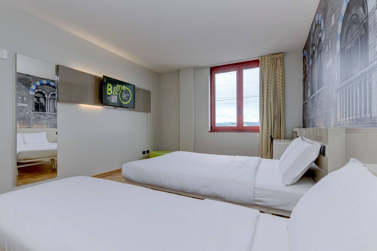 A hotel room with two single beds, a window with red shutters, a wall-mounted TV, and a framed picture.