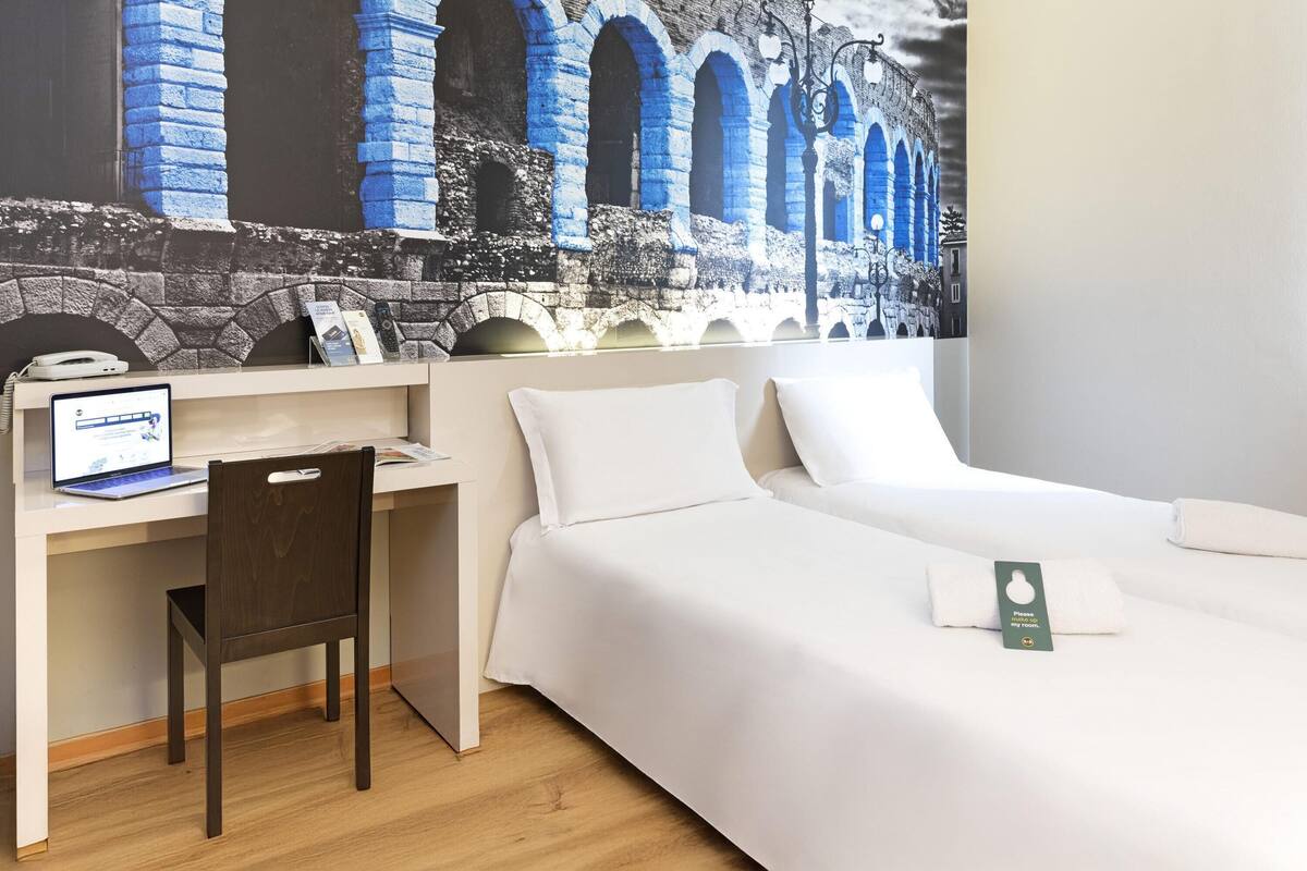A hotel room with a bed, a desk with a laptop, a chair, and a large mural of an ancient aqueduct.