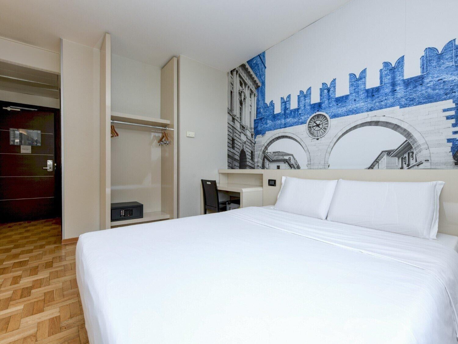 Ein Hotelzimmer mit Bett, Kleiderschrank, Kleiderhaken und Wandgemälde.