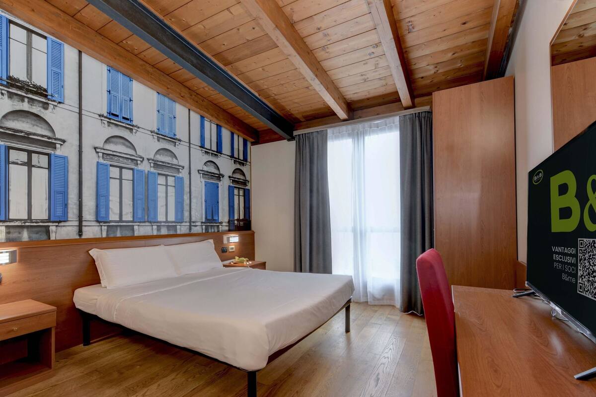 A hotel room with a wooden ceiling, a bed, a desk, and a large window with curtains.