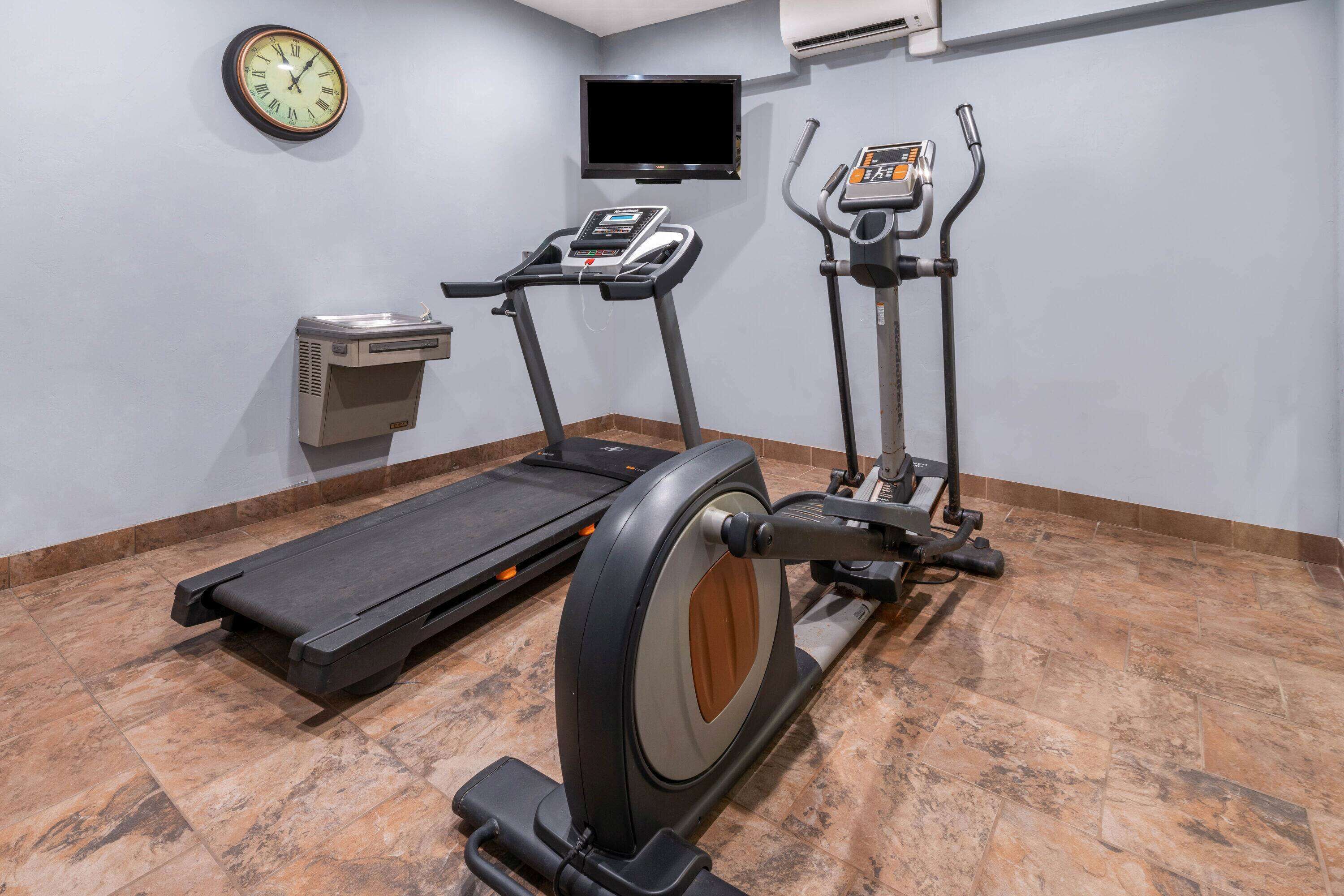 Un gimnasio con cinta de correr, elíptica y bicicleta estática, un reloj y un televisor en la pared.