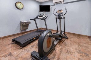 Een sportschool met een loopband, een hometrainer, een vastzetfiets, een klok en een televisie aan de muur.