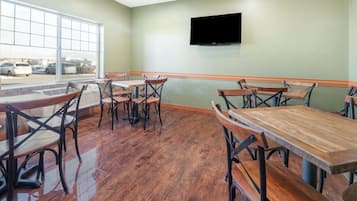 Um restaurante com mesas e cadeiras de madeira, uma janela grande com vista para carros estacionados e uma televisão de tela plana na parede.