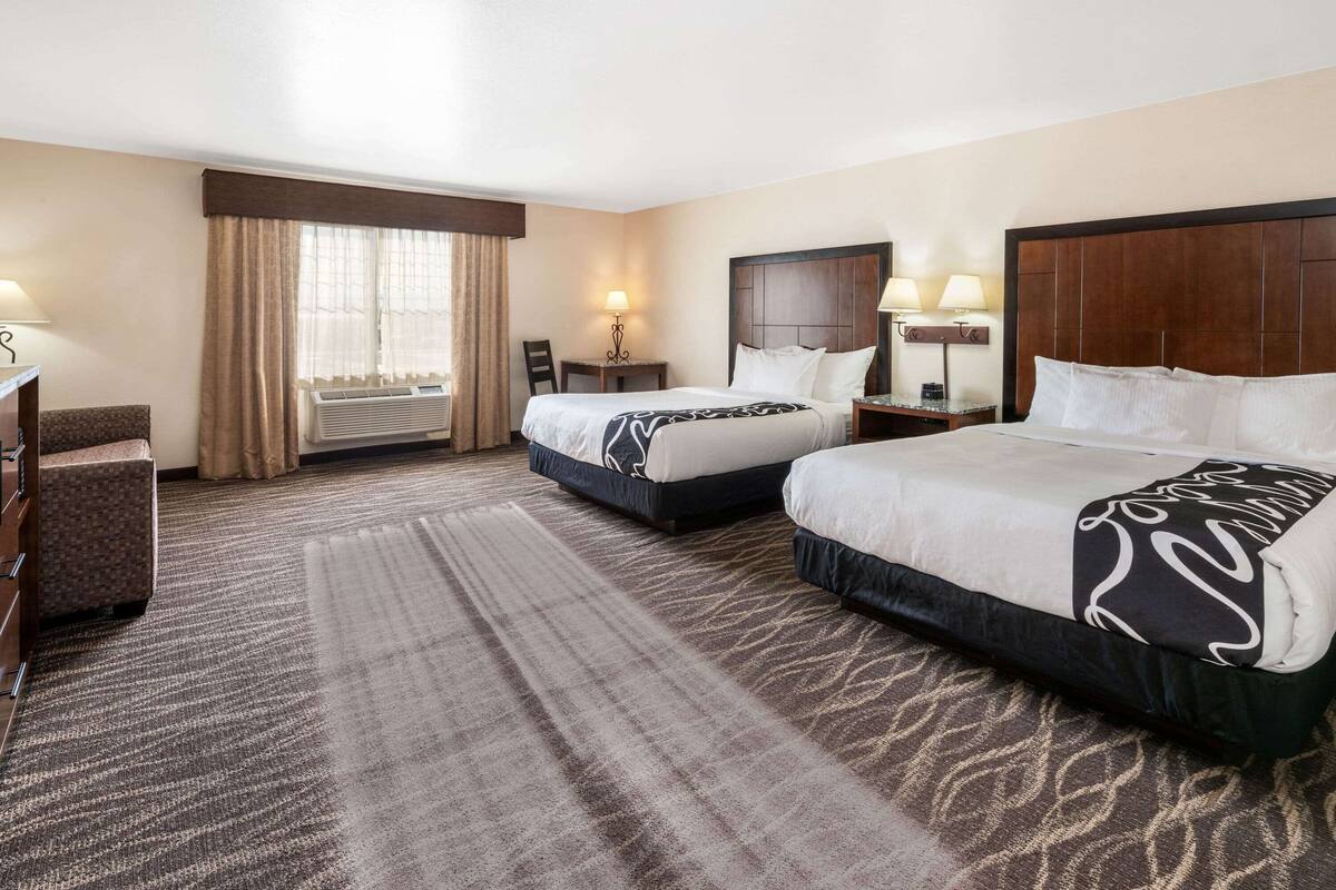 Habitación de hotel con dos camas, un escritorio, una silla y un ventanal con cortinas.