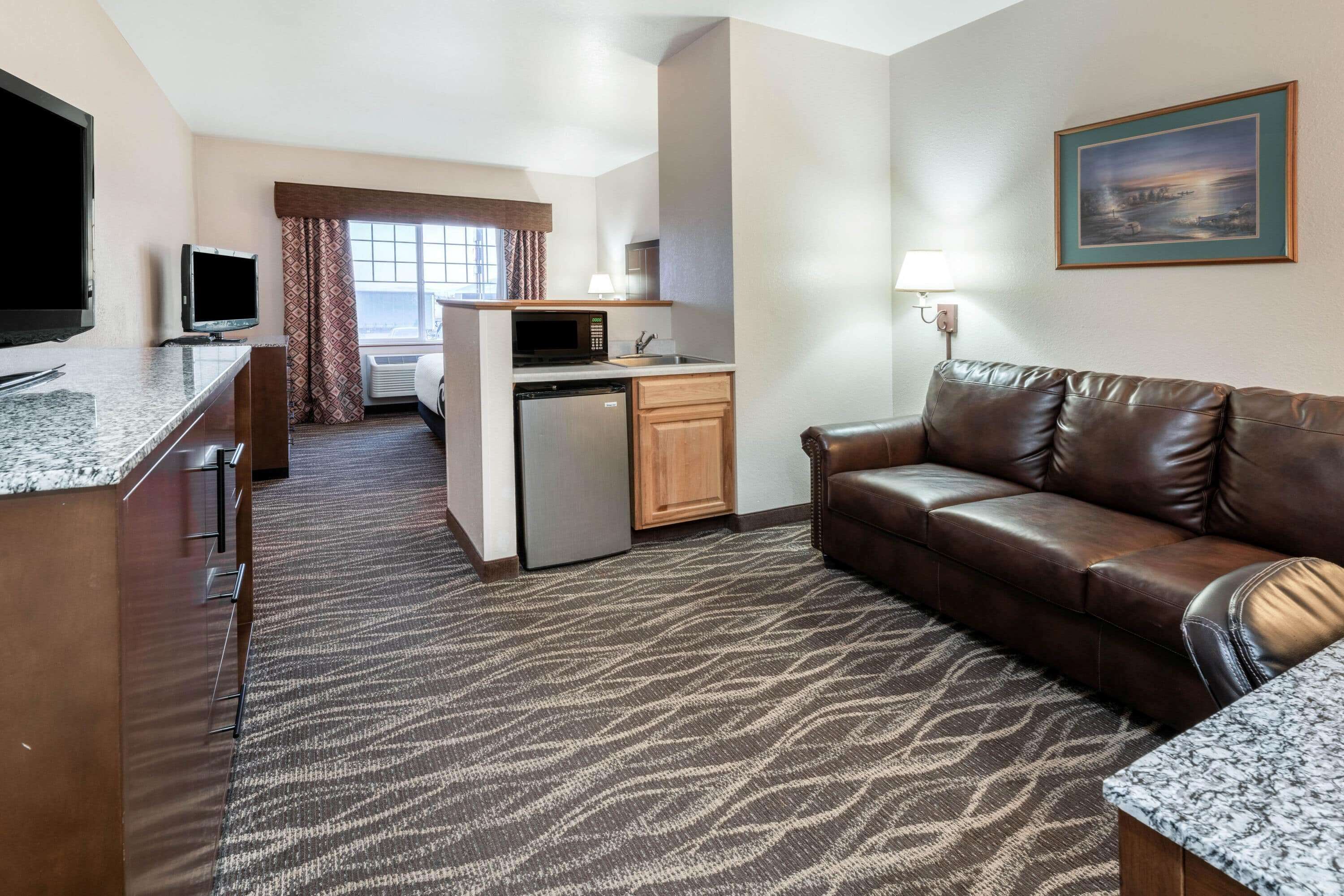 A hotel room with a kitchenette, a leather sofa, a flat-screen TV, and a painting on the wall.