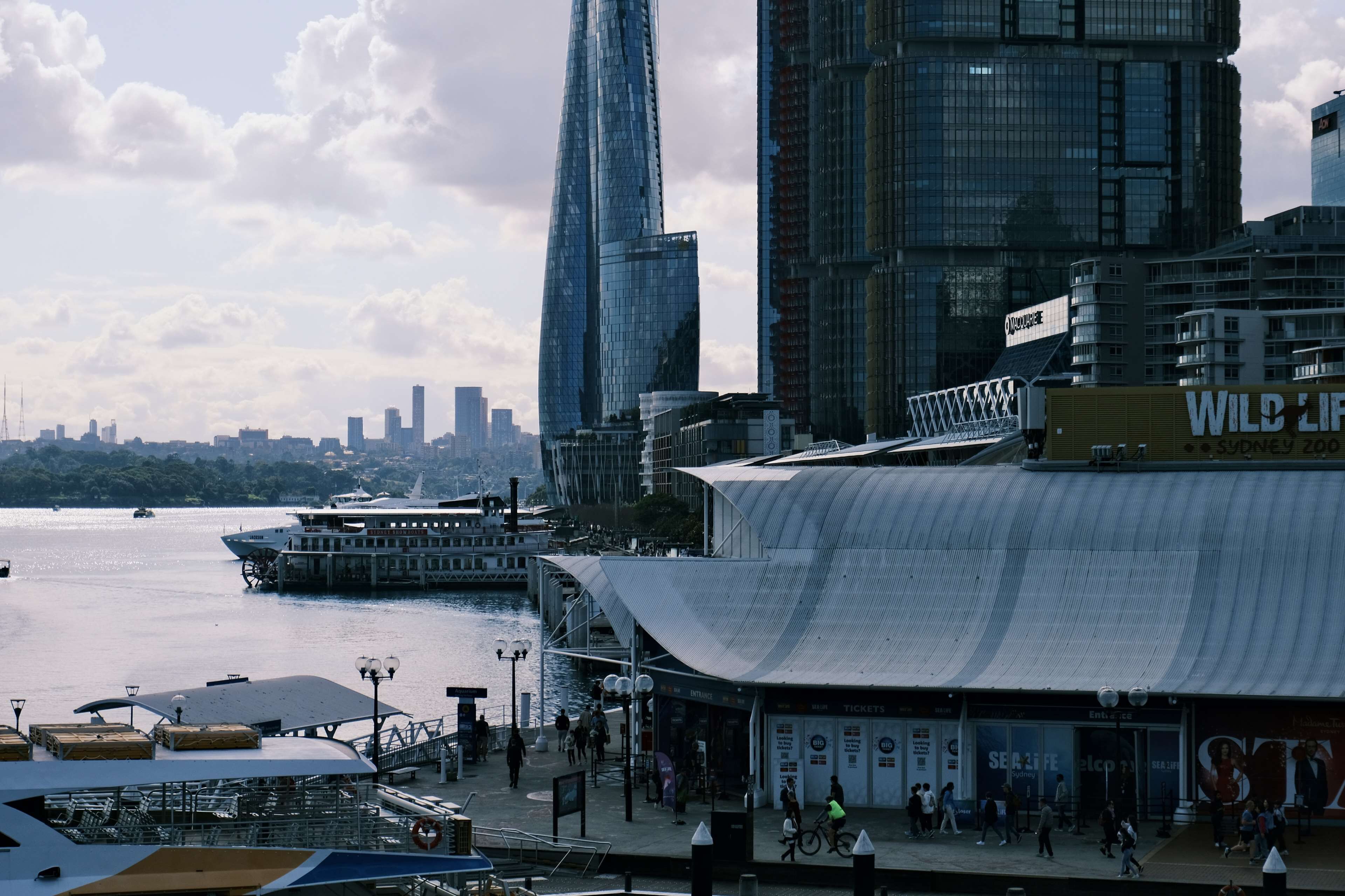 A waterfront promenade with modern buildings, a ferry, and a covered seating area.