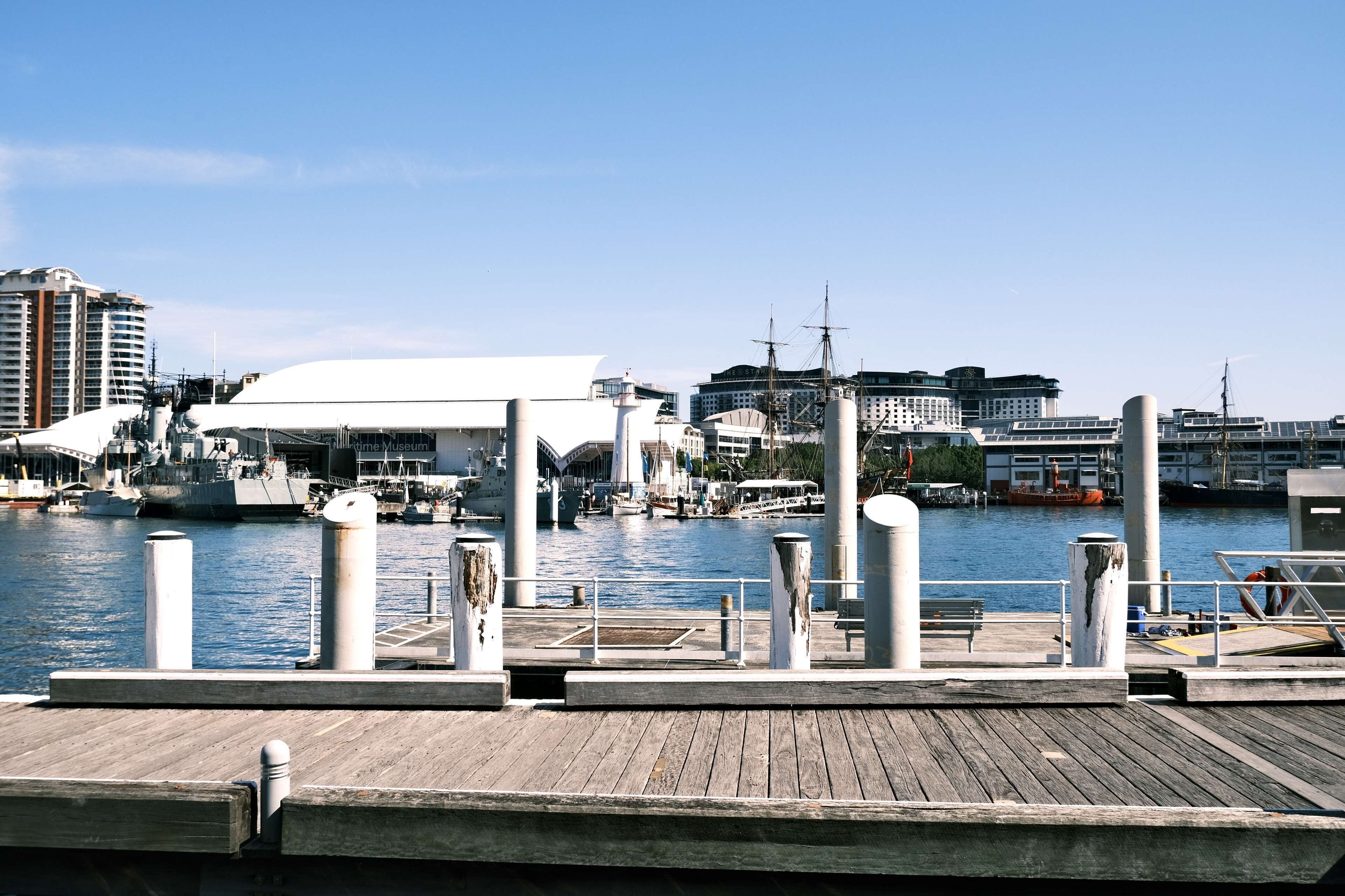 A waterfront dock with a white building, a marina with boats, and a cityscape in the background.