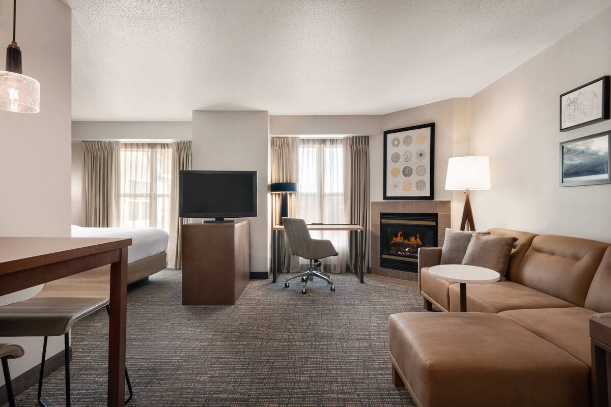 A hotel room with a bed, a desk, a television, a sofa, a chair, a fireplace, and framed artwork on the walls.