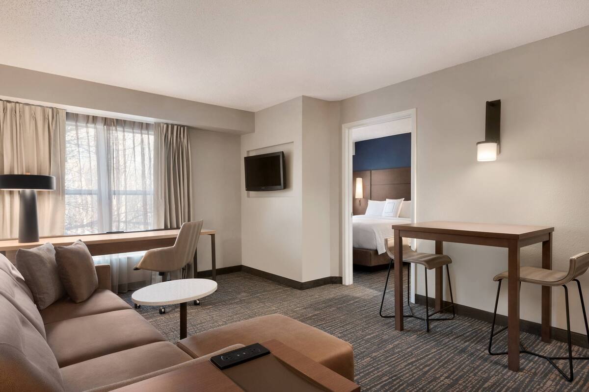 A hotel room with a sofa, a small table, a desk with two chairs, a TV mounted on the wall, and a bed with a headboard.