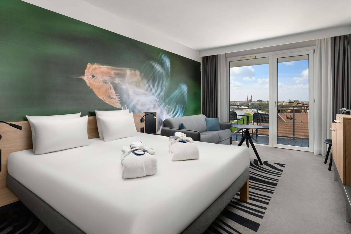 A hotel room with a bed, a desk, a chair, a view of the city, and a balcony.