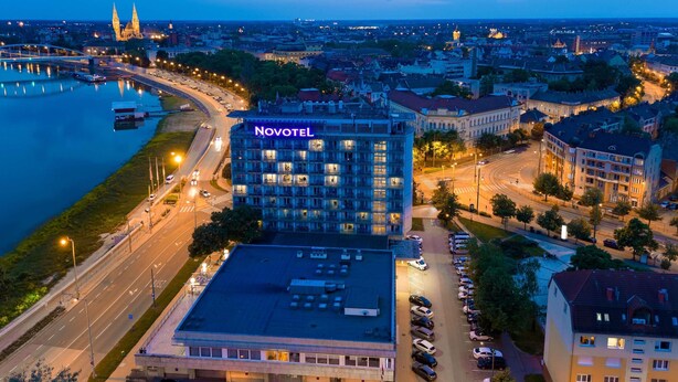 A Novotel hotel with a parking lot, surrounded by city buildings and a river. - Novotel Szeged (Szeged)