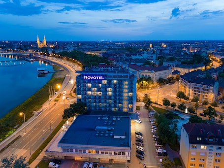 A Novotel hotel with a parking lot, surrounded by a cityscape and a river.. Novotel Szeged