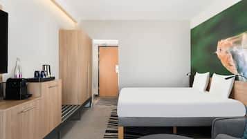 Quarto de hotel moderno com cama, escrivaninha com cafeteira, televisão e um quadro de um pássaro.